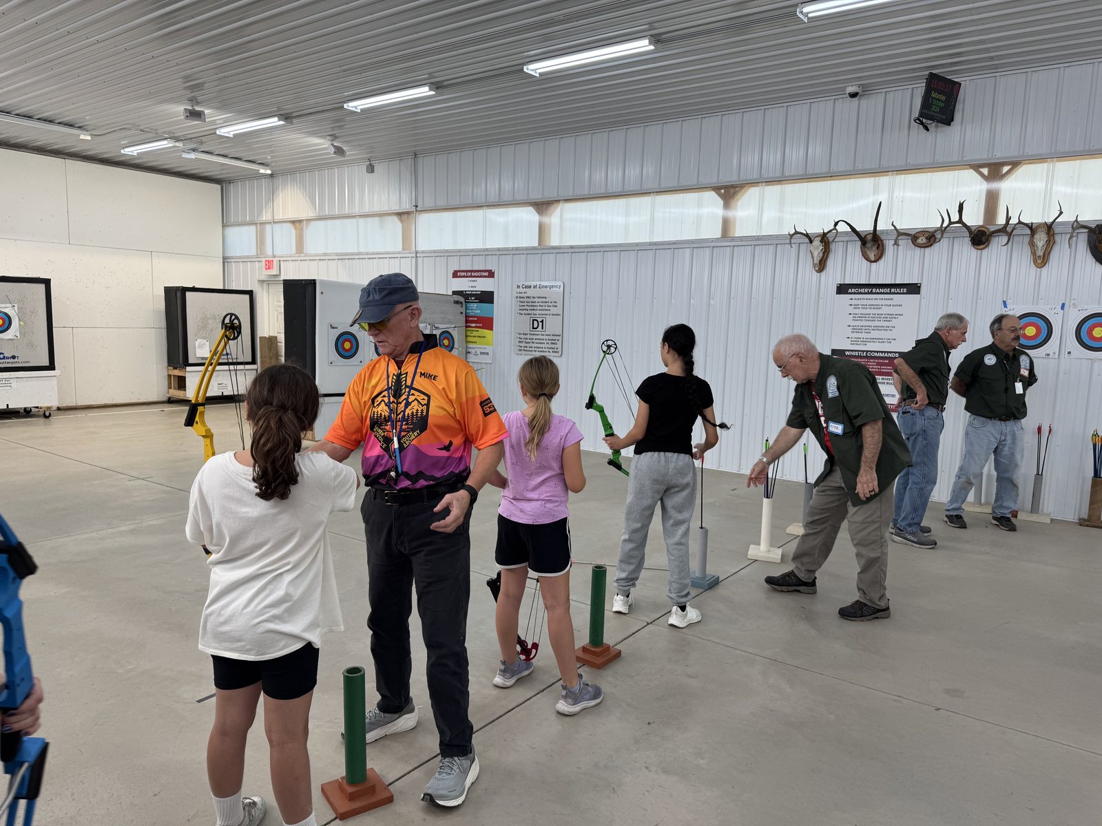 Coaches working one to one with beginner archers on the indoor range, each archer at a ground quiver shooting toward round target faces on the wall.