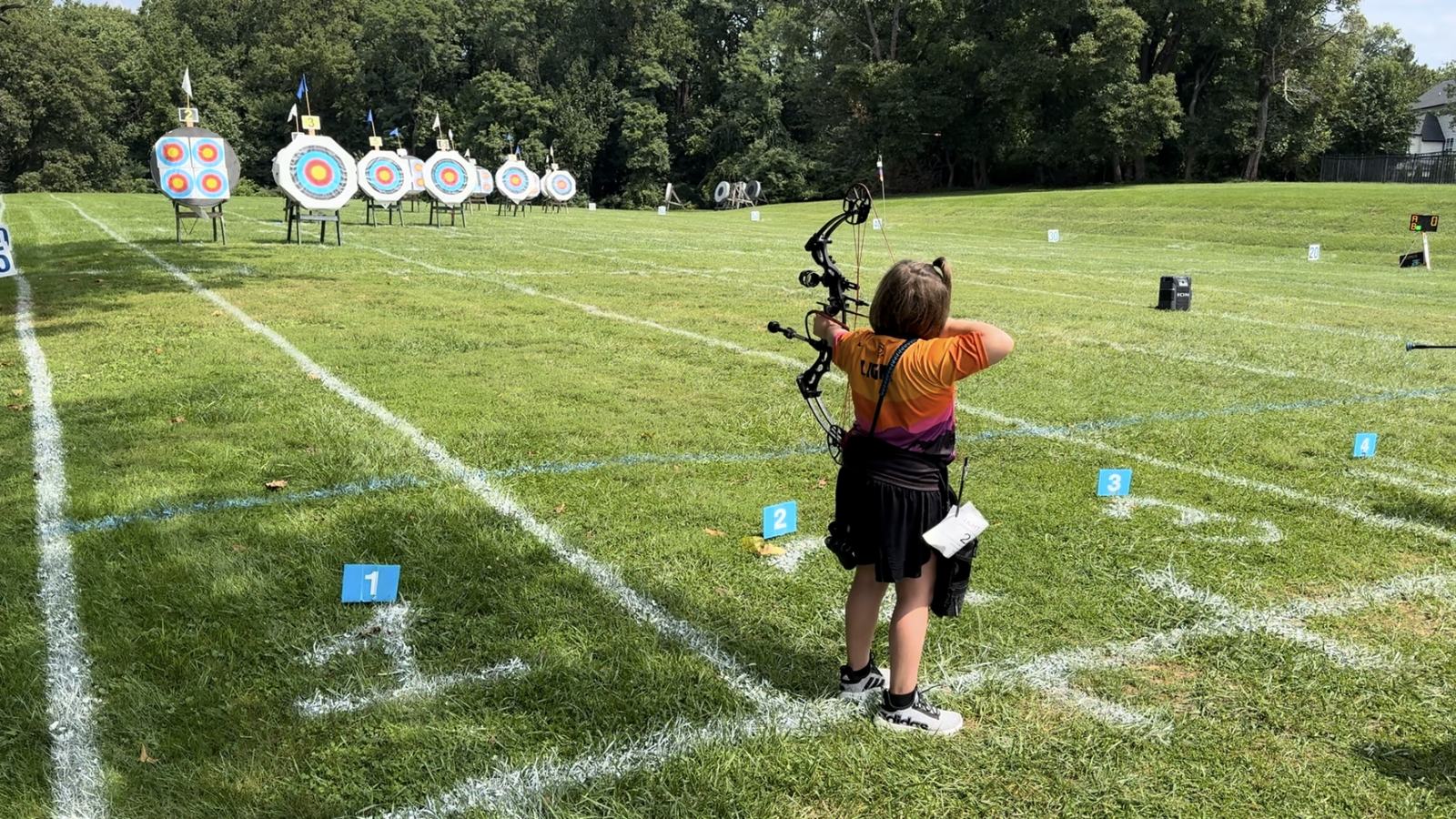 A young archer at full draw on the club's outdoor field range, shooting toward a row of round target faces set out across the grass with numbered distance stakes.