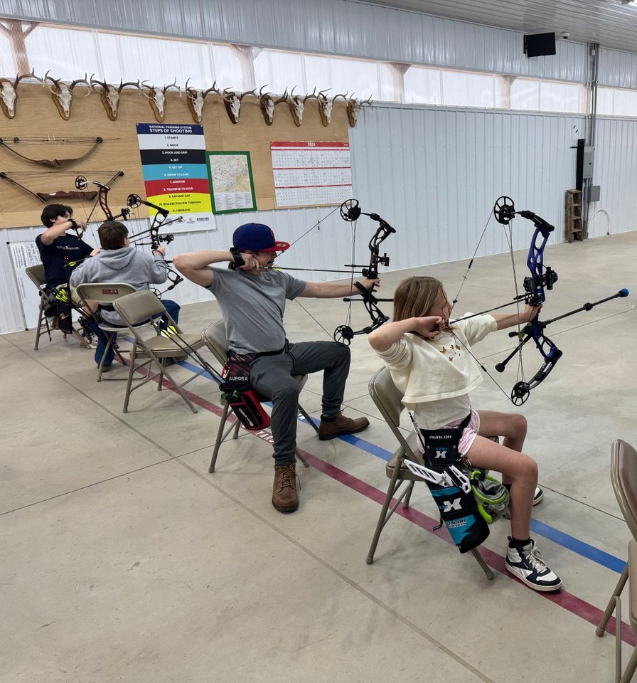 Young archers seated at full draw along the indoor shooting line, drawing compound bows under a coach's eye.