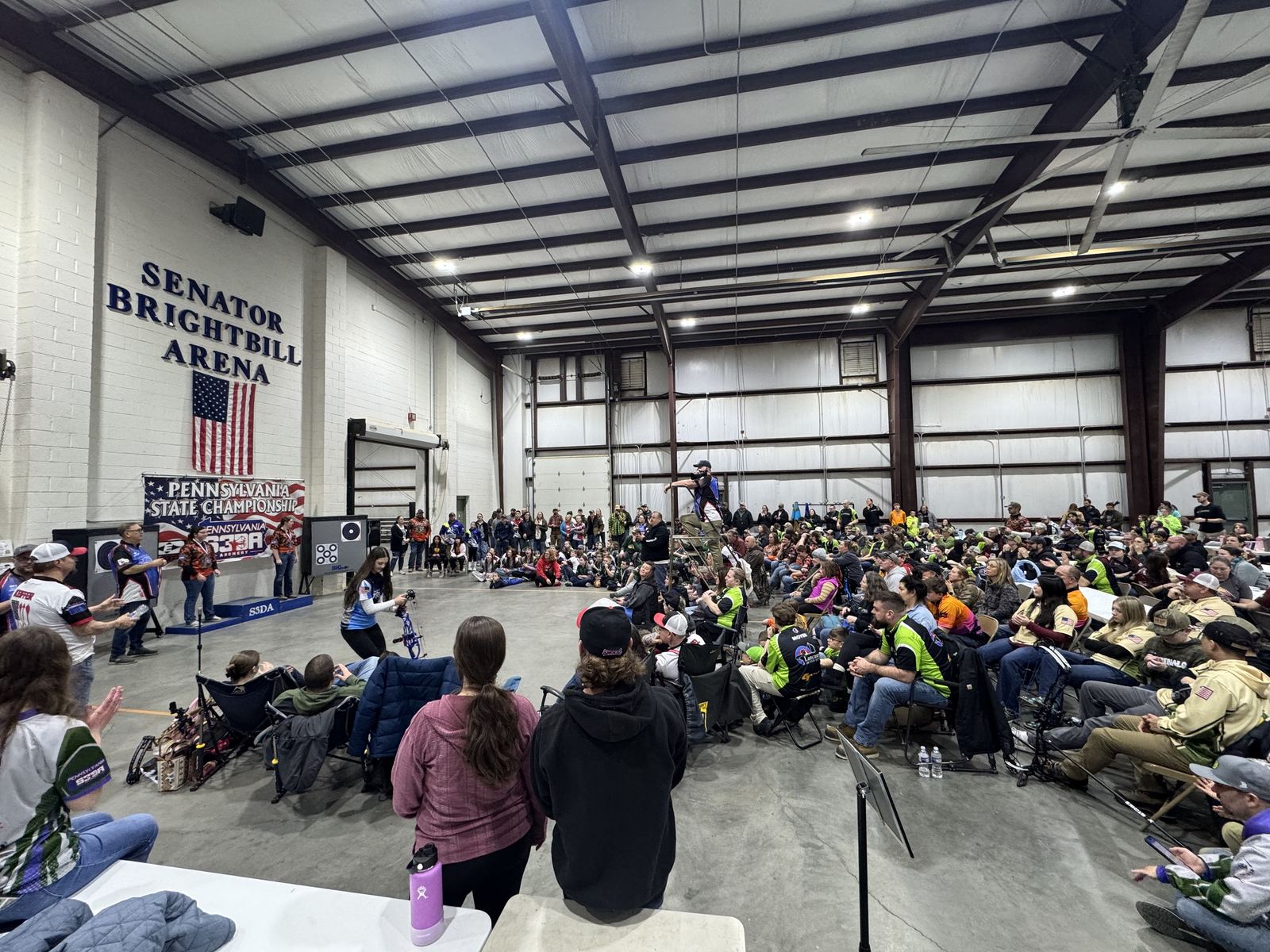 A large indoor arena filled with young archers and families seated for the state championship awards ceremony.
