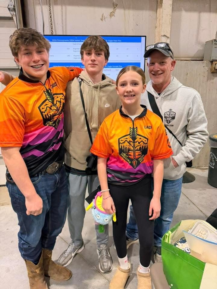 Three young archers in team jerseys with one of their coaches, smiling together at a tournament.